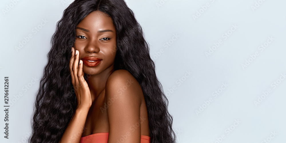 Beautiful African woman portrait. Brunette curly haired young model with dark skin touching her chin and posing against grey background .  - obrazy, fototapety, plakaty 