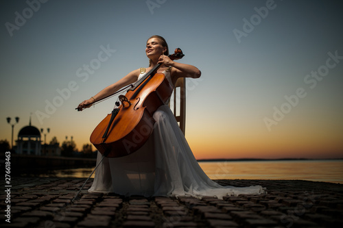 beautiful girl in a white dress playing the cello on the lake after sunset
