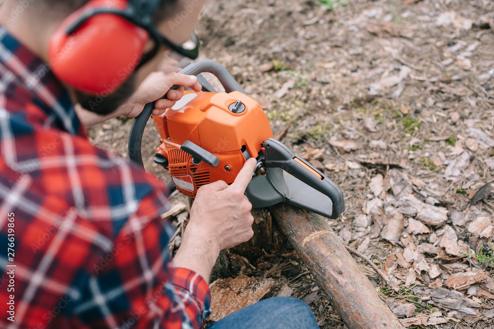 selective focus of lumberjack in hearing protectors repairing chainsaw in forest selective focus of lumberjack in hearing protectors repairing chainsaw in forest