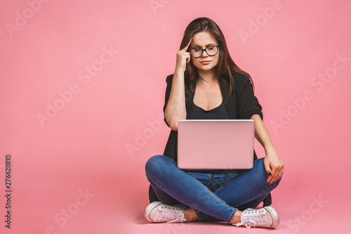 Angry sad bored woman in stress sitting on the floor with laptop computer isolated against pink background.