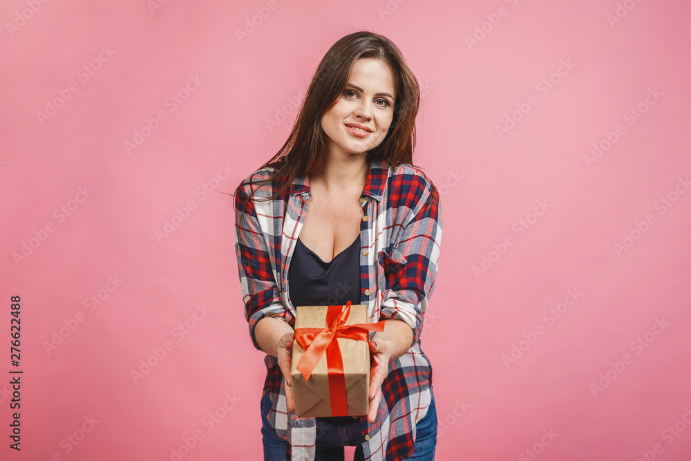 Happy brunette woman in casual holding gift box and looking at the camera while enjoys over pink background Happy brunette woman in casual holding gift box and looking at the camera while enjoys over pink background