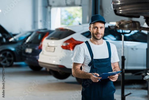 happy bearded car mechanic looking at camera and holding clipboard near cars photo
