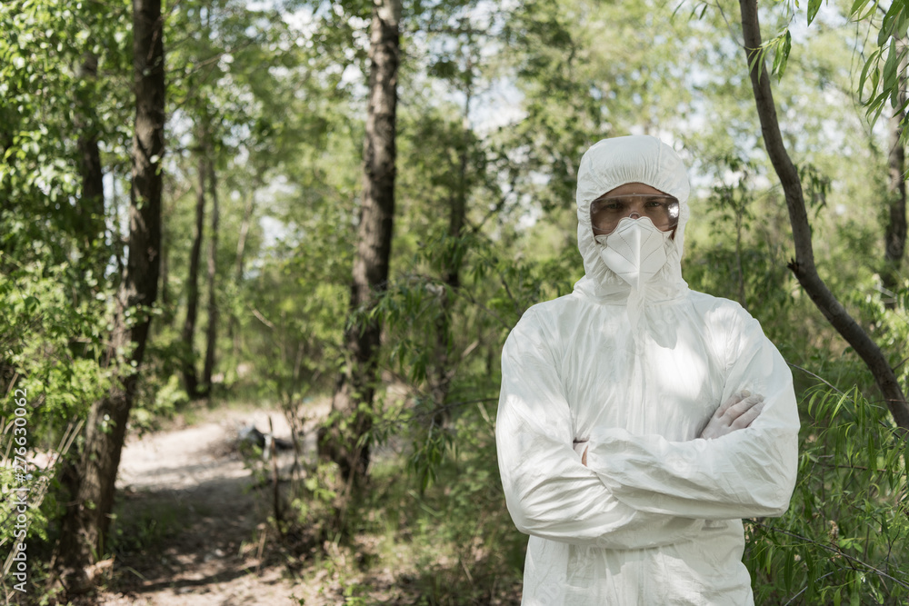 front view of ecologist in protective costume and respirator with crossed arms in forest front view of ecologist in protective costume and respirator with crossed arms in forest