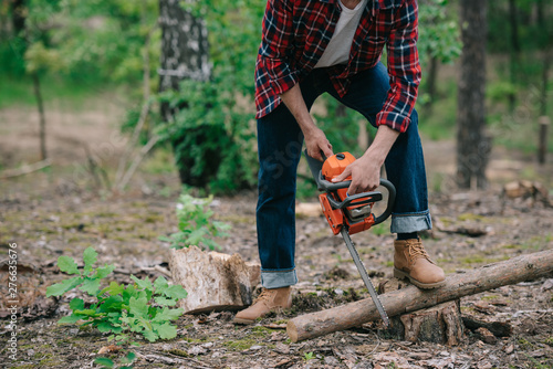 cropped view of lumberman in plaid shirt and denim jeans cutting wood with chainsaw in forest