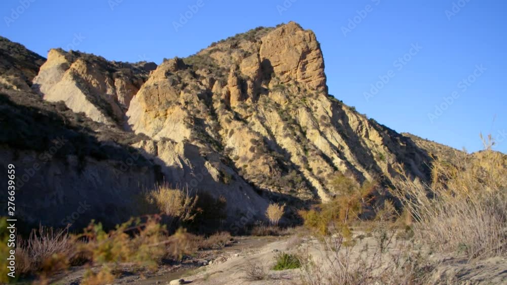 Badlands mountain near a dry river. Located in de Almería desert, the only desert in Europe. Desertic vegetation, warm color of the terrain. Blue sky in the background Badlands mountain near a dry river. Located in de Almería desert, the only desert in Europe. Desertic vegetation, warm color of the terrain. Blue sky in the background