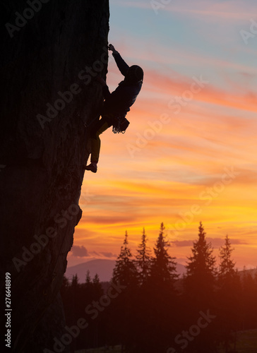 Male silhouette of climber climbing at nightfall and reaching for his next hold hand. Sunset orange cloudy sky and mountain landscape on background. Snapshot with low angle view and copy space
