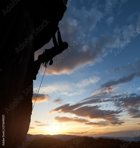 Athletic man silhouette climbing in safety harness on overhanging cliff rock high up over mountains with sunrise sky on background. Copy space. Side view. Success, leadership, chievements concept