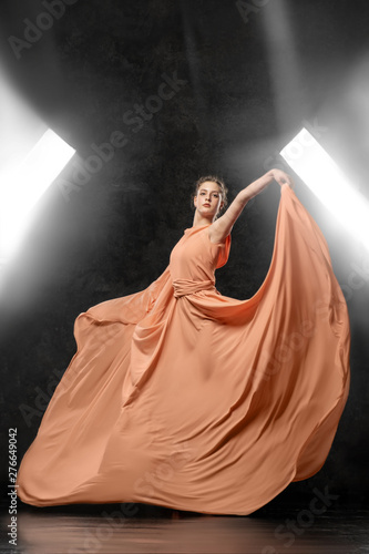 Ballerina. A young dancer dressed in a long peach dress, pointe shoes with ribbons. Performs a graceful, graceful dance movement on the background of the bright light of the window.  Ballet studio. 3D