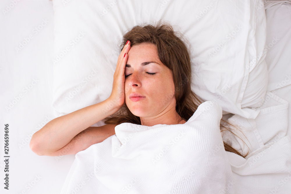 A young woman holds her head and lies in bed. Concept insomnia, headache, malaise, illness. Flat lay, top view A young woman holds her head and lies in bed. Concept insomnia, headache, malaise, illness. Flat lay, top view