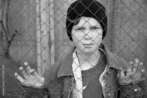 Homeless little boy standing behind mesh outdoors