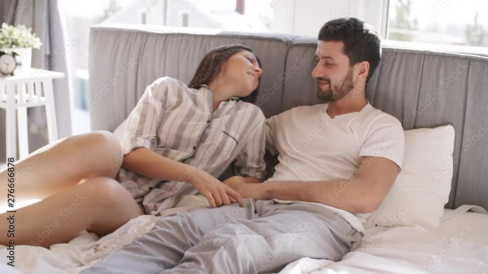 Handheld shot of young woman in pajama lying down on bed next to her boyfriend and kissing him on lips. Couple relaxing together in morning Handheld shot of young woman in pajama lying down on bed next to her boyfriend and kissing him on lips. Couple relaxing together in morning