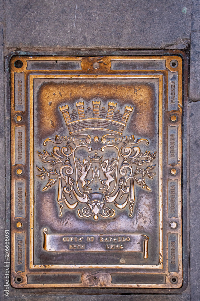 Close-up of a golden cast iron manhole cover in a street pavement with the symbols of the municipal coat of the city of Rapallo, rampant griffons holding the Marian acronym, Genoa, Liguria, Italy