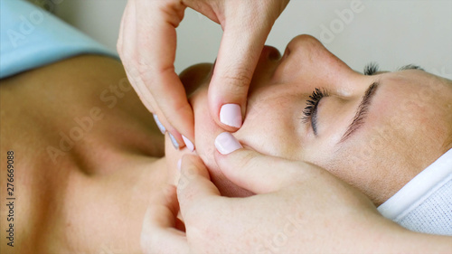 Young woman on healthy massage treatment in cosmetology spa center. Massage therapist is doing manual massage on client's face.