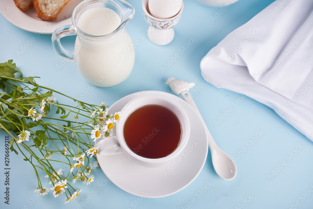 Morning. Breakfast. Cup of tea with milk, jar with milk, egg on the blue background Morning. Breakfast. Cup of tea with milk, jar with milk, egg on the blue background
