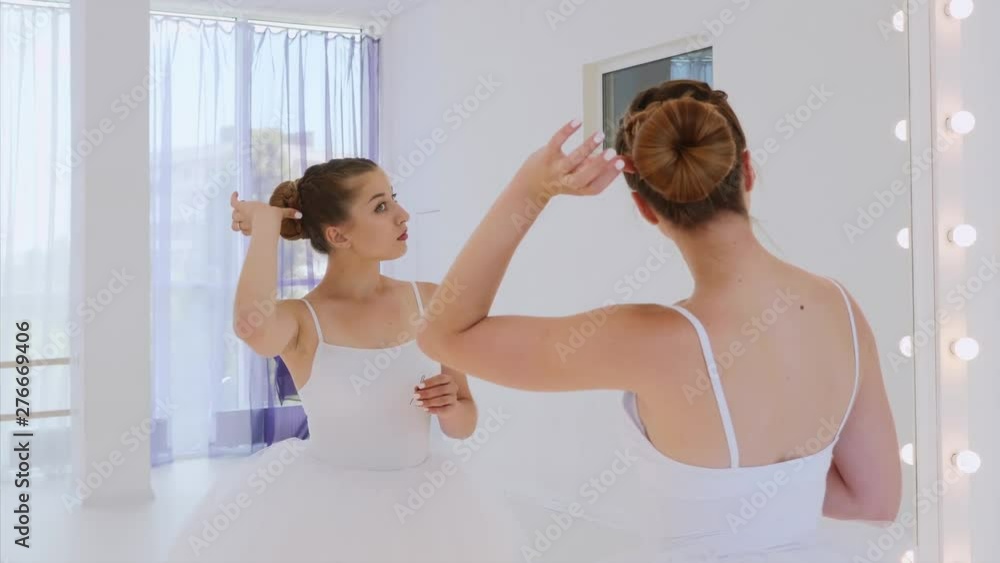 Ballerina in white tutu does her hair standing in front of the mirror in theatre dance class. She fixes the hair pulled into a bun with bobby pins. Portrait shot in mirror reflection. Ballerina in white tutu does her hair standing in front of the mirror in theatre dance class. She fixes the hair pulled into a bun with bobby pins. Portrait shot in mirror reflection.