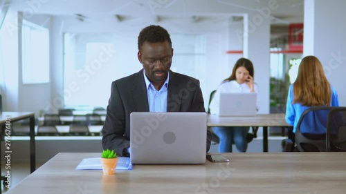 Mature man sitting at desk and typing in his laptop. He is very concentated on work. Then he looks into the camera. His colleagues are working on background photo