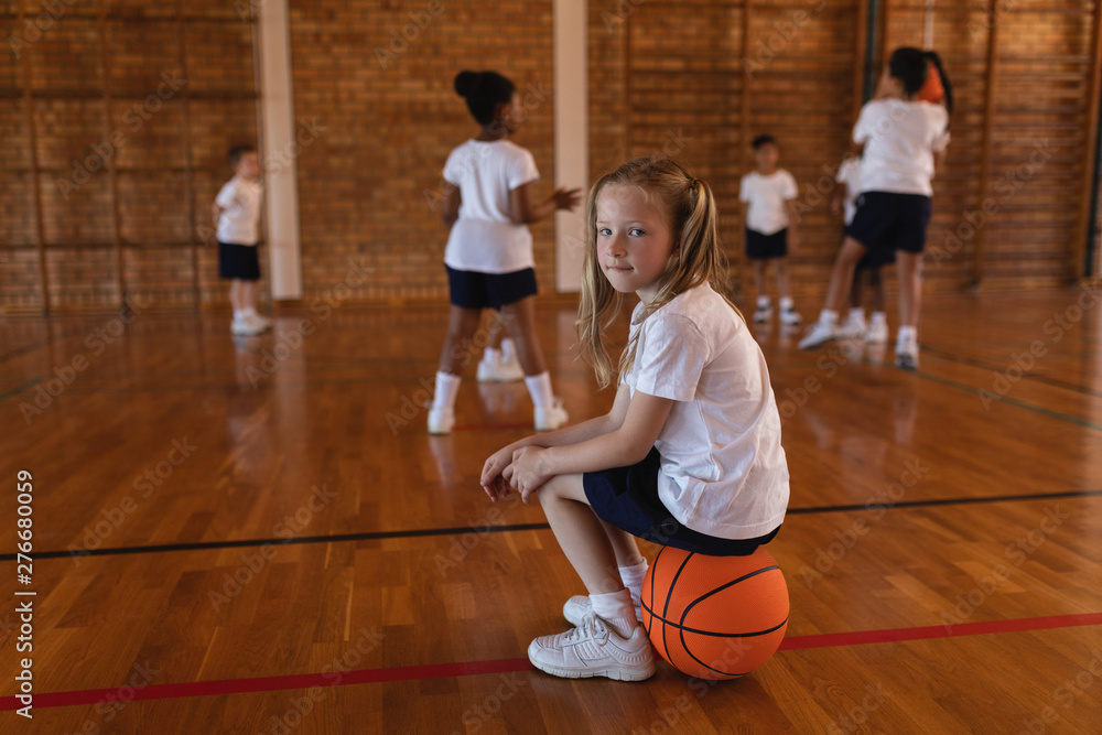 Side view of schoolgirl sitting on basketball and looking at camera at basketball court
