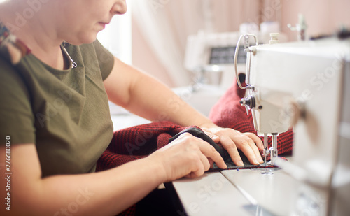 Woman clothier sitting at workplace in atelier stitching red skirt with professional sewing machine. Seamstress hand pulling fabric under presser foot. Handmade, hobby, business concept. Close up view photo