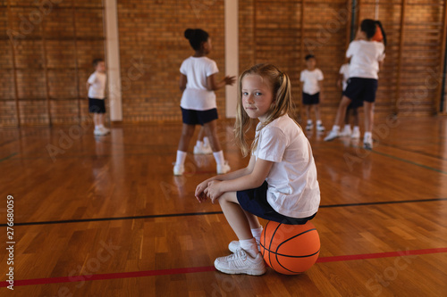 Side view of schoolgirl sitting on basketball and looking at camera at basketball court