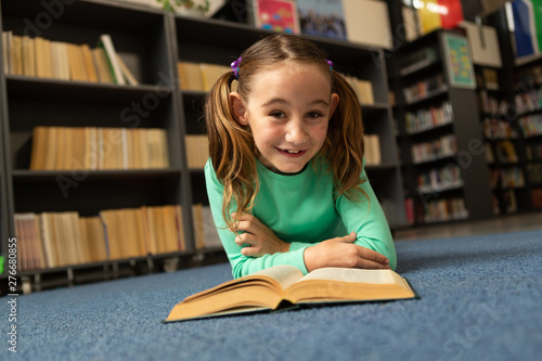 Smiling schoolgirl with book lying on floor and looking at camera in library