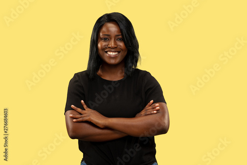 Young african-american woman isolated on yellow studio background, facial expression. Beautiful female half-length portrait. Concept of human emotions, facial expression. Standing crossing hands. © master1305