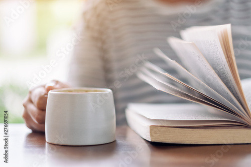 Closeup image of a woman holding and reading a book while drinking coffee on wooden table