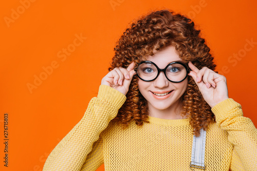 curly- haired woman ajusting her eyeglasses on orange background. tennage girl with stylish hipster glasses photo