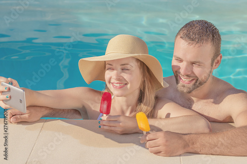 Summertime in pool. Young beautiful couple with ice-cream taking selfie in the swimming pool.