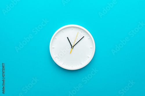 White wall clock with yellow second hand hanging on the wall. Minimalist flat lay image of plastic wall clock over blue turquiose background with copy space and central composition.