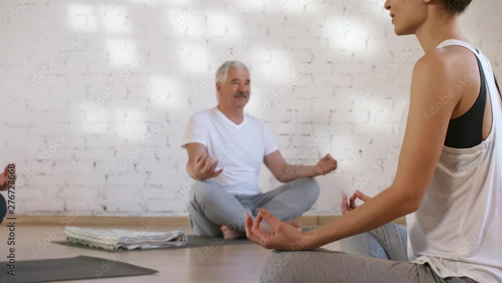 Panning shot of young female yoga teacher giving class for middle-aged Caucasian couple, sitting with crossed legs on mats, and instructing them to make mudras with hands on top of knees and relax Panning shot of young female yoga teacher giving class for middle-aged Caucasian couple, sitting with crossed legs on mats, and instructing them to make mudras with hands on top of knees and relax