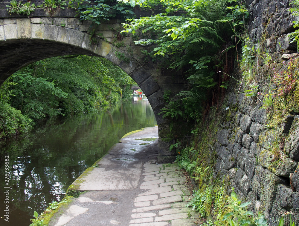 a path along the rochdale canal crossing under an old stone bridge  overgrown with vegetation with trees and a narrowboat in the distance  Photos | Adobe Stock