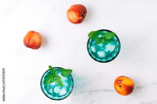 Two beautiful vintage turquoise glasses with cold drink and ice cubes, decorated with fresh green mint leaves and three nectarins on white marble table. Top view. photo