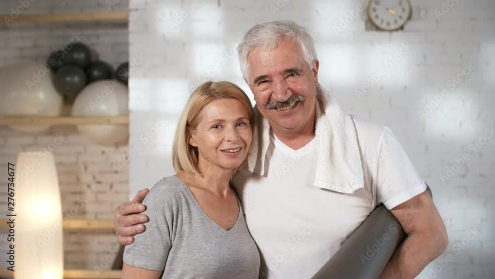 Waist-up portrait shot of happy mature Caucasian husband and wife, wearing t-shirts and holding rolled mat, standing in their own loft-style yoga studio, hugging affectionately and smiling for camera Waist-up portrait shot of happy mature Caucasian husband and wife, wearing t-shirts and holding rolled mat, standing in their own loft-style yoga studio, hugging affectionately and smiling for camera