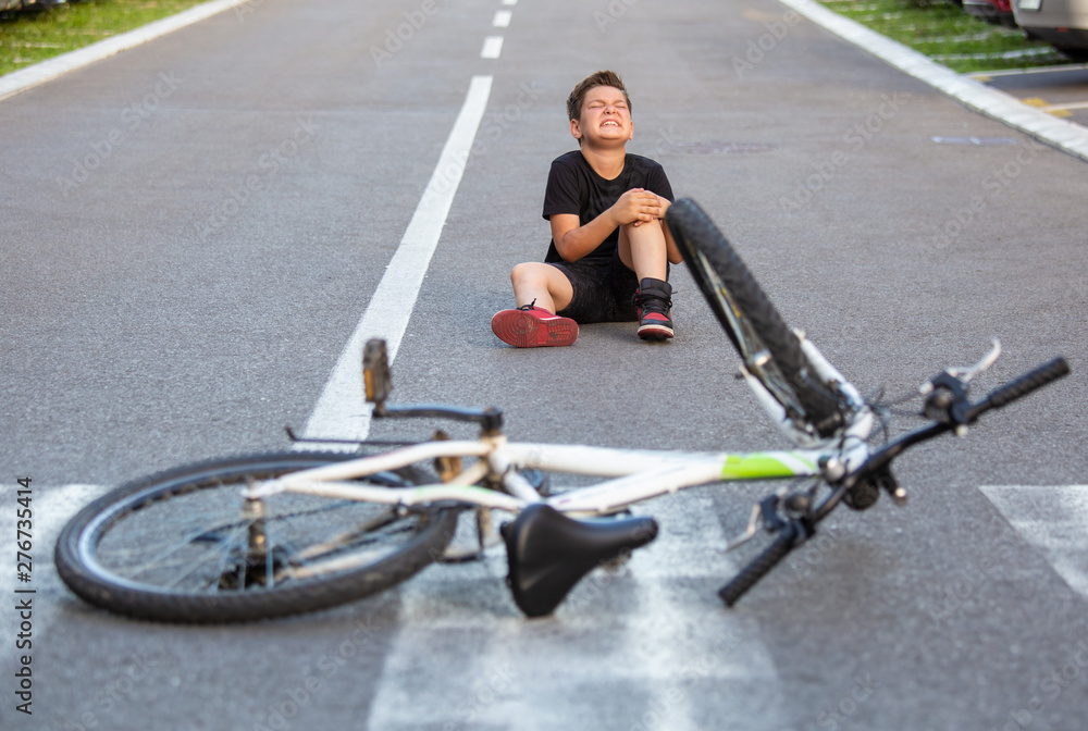 Kid hurts his leg after falling off his bicycle. Child is learning to ride a bike. Boy in the street ground with a knee injury screaming after falling off to his bicycle. - obrazy, fototapety, plakaty 