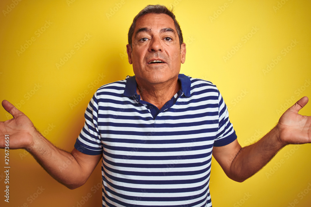 Handsome middle age man wearing striped polo standing over isolated yellow background clueless and confused expression with arms and hands raised. Doubt concept. Handsome middle age man wearing striped polo standing over isolated yellow background clueless and confused expression with arms and hands raised. Doubt concept.