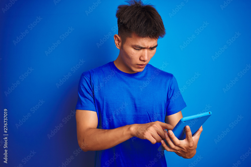 Young asian chinese man using tablet standing over isolated blue background scared in shock with a surprise face, afraid and excited with fear expression Young asian chinese man using tablet standing over isolated blue background scared in shock with a surprise face, afraid and excited with fear expression