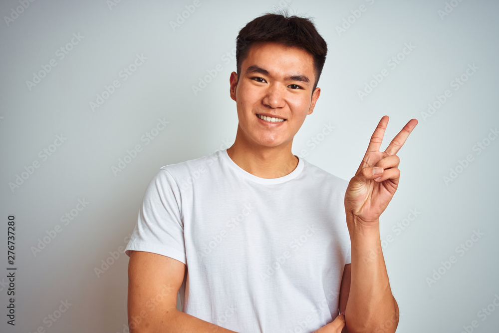 Young asian chinese man wearing t-shirt standing over isolated white background smiling with happy face winking at the camera doing victory sign with fingers. Number two. Young asian chinese man wearing t-shirt standing over isolated white background smiling with happy face winking at the camera doing victory sign with fingers. Number two.