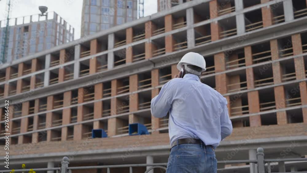 Profession, business and telecommunications, architect talking on cell phone and walking in construction site under building scaffolding. Steadicam shot 4K Profession, business and telecommunications, architect talking on cell phone and walking in construction site under building scaffolding. Steadicam shot 4K