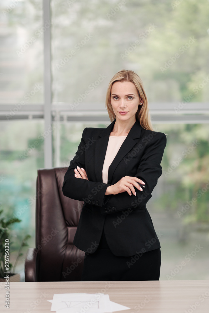 Young attractive Caucasian blond woman in black business suit sits at desk in bright office. Studying paper documents, looking at camera and smiling. Young attractive Caucasian blond woman in black business suit sits at desk in bright office. Studying paper documents, looking at camera and smiling.