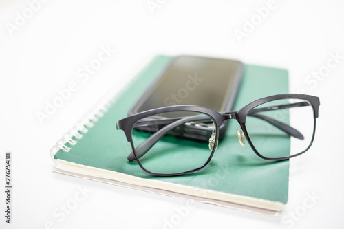 Close up of reading glasses on mobile smartphone and green notebook on white table. Education technology and work concept.
