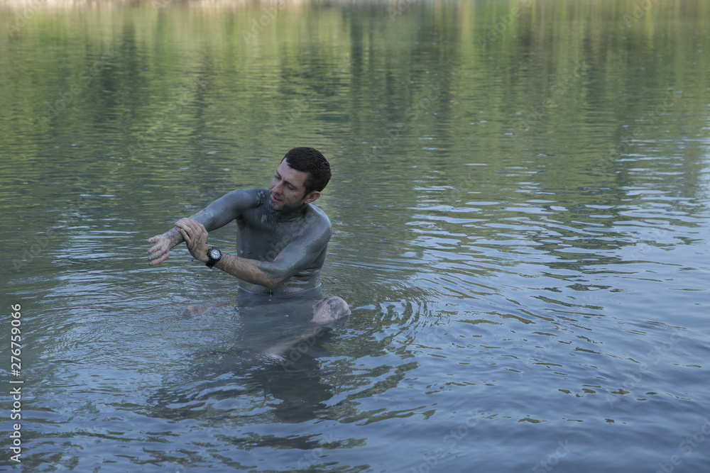 A young handsome shirtless man, his body smeared with mud, sits and washes himself in the water of a mountain river. Healthy lifestyle, natural procedures. The concept of unity with nature. A young handsome shirtless man, his body smeared with mud, sits and washes himself in the water of a mountain river. Healthy lifestyle, natural procedures. The concept of unity with nature.