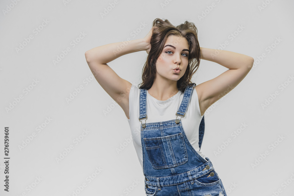 A teenage girl with pleasure an attractive look. Wears fashionable denim overalls and white shirt. Having put his hands on his head, he looks at the camera. The concept of people, style and fashion. A teenage girl with pleasure an attractive look. Wears fashionable denim overalls and white shirt. Having put his hands on his head, he looks at the camera. The concept of people, style and fashion.