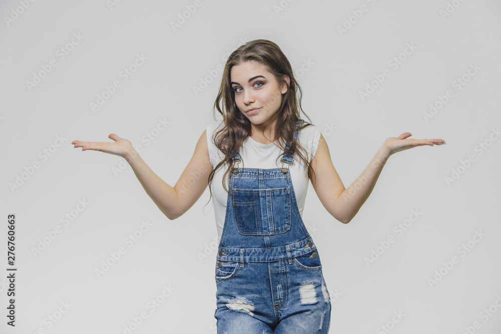 Happy brunette teenager standing on gray background. Dressed in a denim overalls and a white T-shirt. Looking at the camera, the girl put her hands in different directions, surprisingly. Happy brunette teenager standing on gray background. Dressed in a denim overalls and a white T-shirt. Looking at the camera, the girl put her hands in different directions, surprisingly.