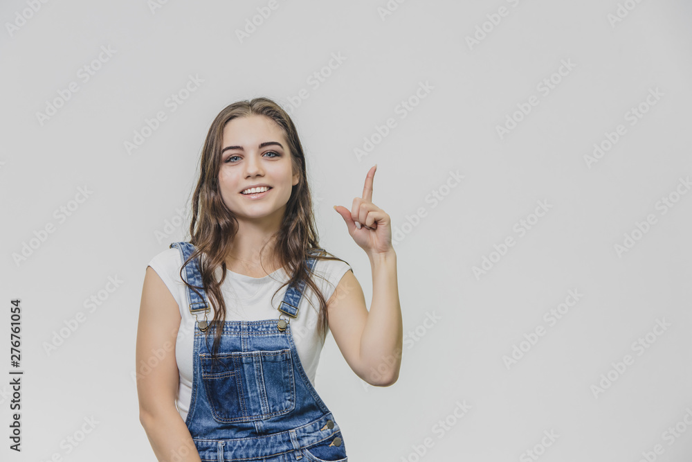 Young attractive student girl shows her index finger up. Tries to concentrate and looks into the camera. Dressed in white t-shirt and denim overalls. Young attractive student girl shows her index finger up. Tries to concentrate and looks into the camera. Dressed in white t-shirt and denim overalls.