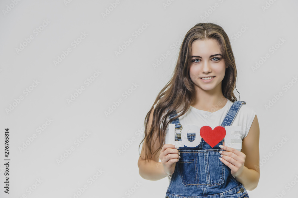 A young beautiful girl is on a gray background. Dressed in a denim suit and a white T-shirt. Holds a sign with a word of love. Opening his blue eyes looks into the camera. A young beautiful girl is on a gray background. Dressed in a denim suit and a white T-shirt. Holds a sign with a word of love. Opening his blue eyes looks into the camera.