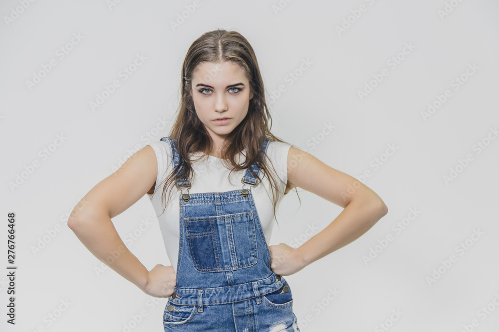 A young beautiful girl stands on a gray background. It is dressed in a denim suit and a white T-shirt. During this, he holds his arms at his waist and looks at the camera. A young beautiful girl stands on a gray background. It is dressed in a denim suit and a white T-shirt. During this, he holds his arms at his waist and looks at the camera.