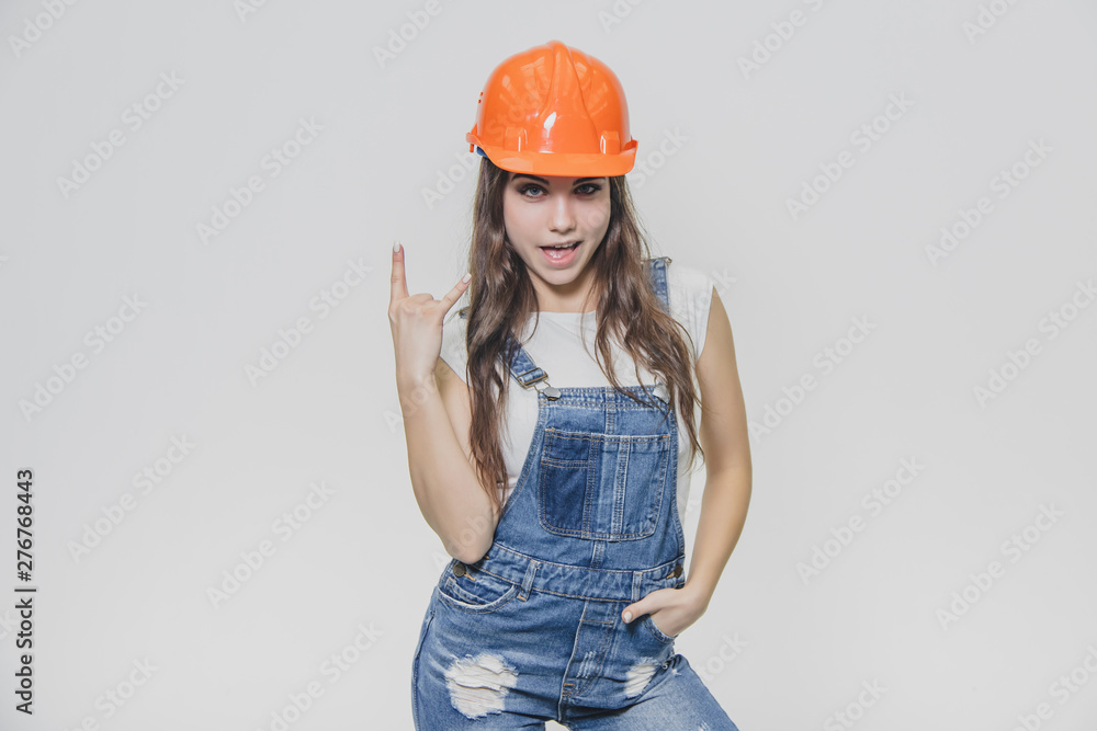 Young pretty girl standing on a white background. During this time wearing a white T-shirt and denim overalls. Dressed in an orange helmet.