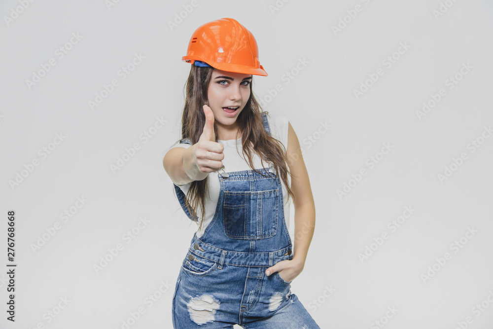 Young pretty girl standing on a white background. During this time wearing a white T-shirt and denim overalls. Dressed in an orange helmet. Young pretty girl standing on a white background. During this time wearing a white T-shirt and denim overalls. Dressed in an orange helmet.