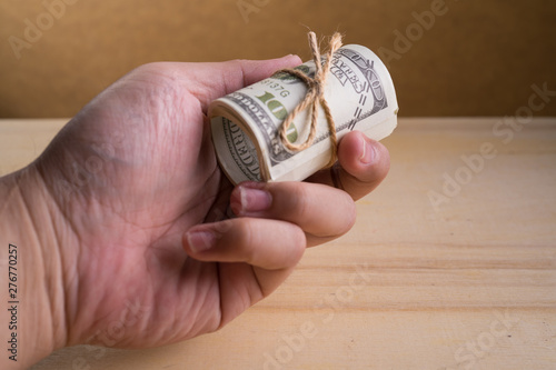 A man Hand and a roll of American currency (USD, American dollars) with 100 dollars bank notes on the outside as a symbol of plenty of money on the wooden background. 