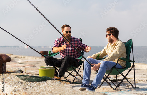 leisure and people concept - happy male friends with rods on pier at sea telling stories about fishing